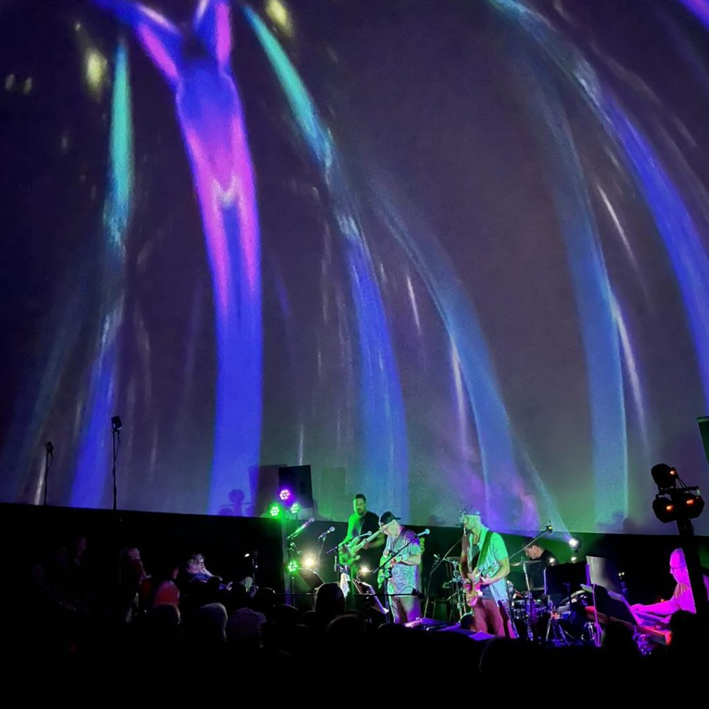 band playing music in a planetarium under graphic images projected on the dome above