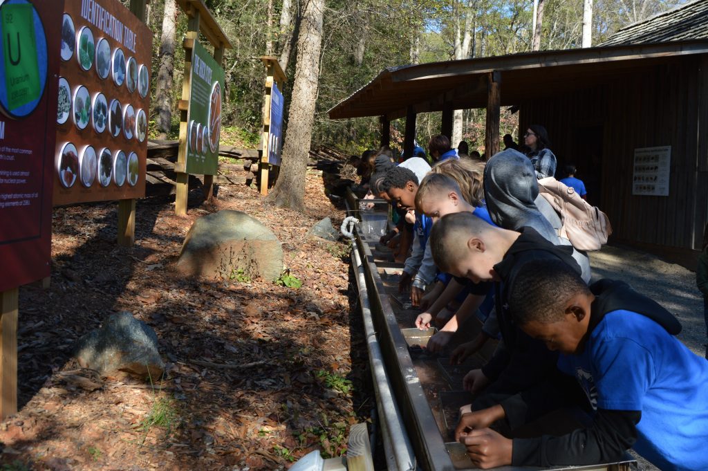 Photograph of a group of kids doing the Gemstone Mining in front of the Grist Mill, sleuthing out the sand from their trays to find gems.