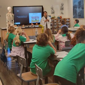 An educator wearing a lab coat stands in front of a group of students. She is accompanied by a human skeleton.