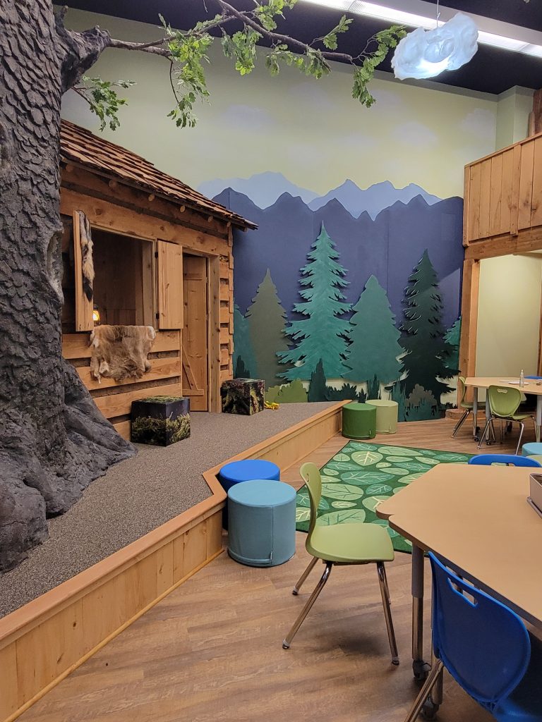 Indoor room with a fake tree framing the left side, a wood house protruding from behind the tree, and a nice mountain-scape painted on the wall. There are tables and chairs framing the right side of the photo. There are small cylinder stools scattered across the floor. There is a stage in this room where the house sits.
