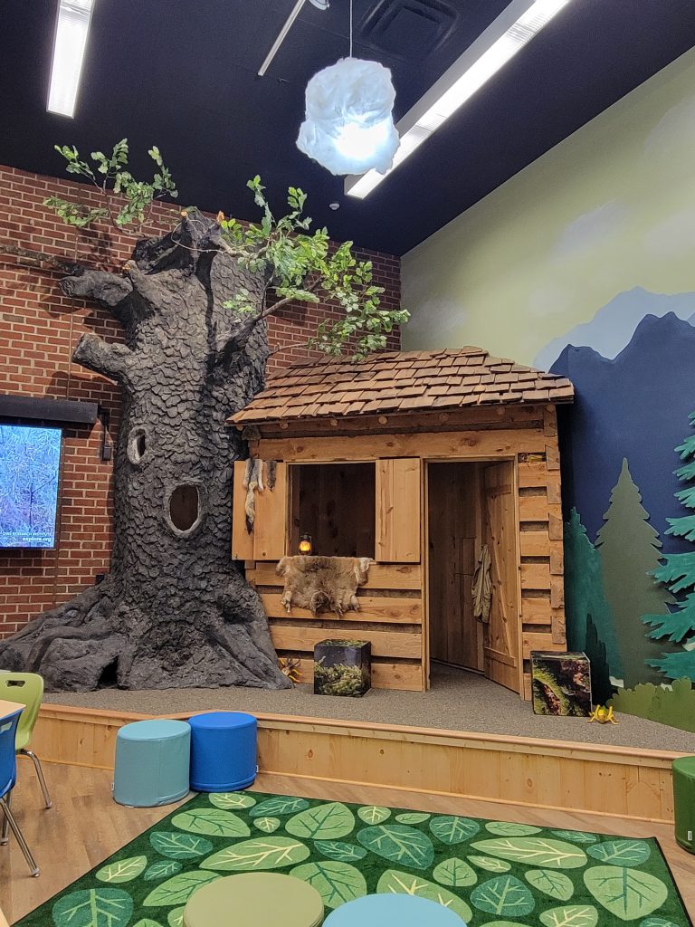 wood/log cabin built in to the corner of the room displayed. There is a replica of a tree beside the house, with a small window to look out of. There is a rug with leaf patterns on the floor beneath the stage.