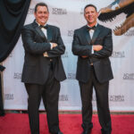 Two men posing in black suits in front of backdrop